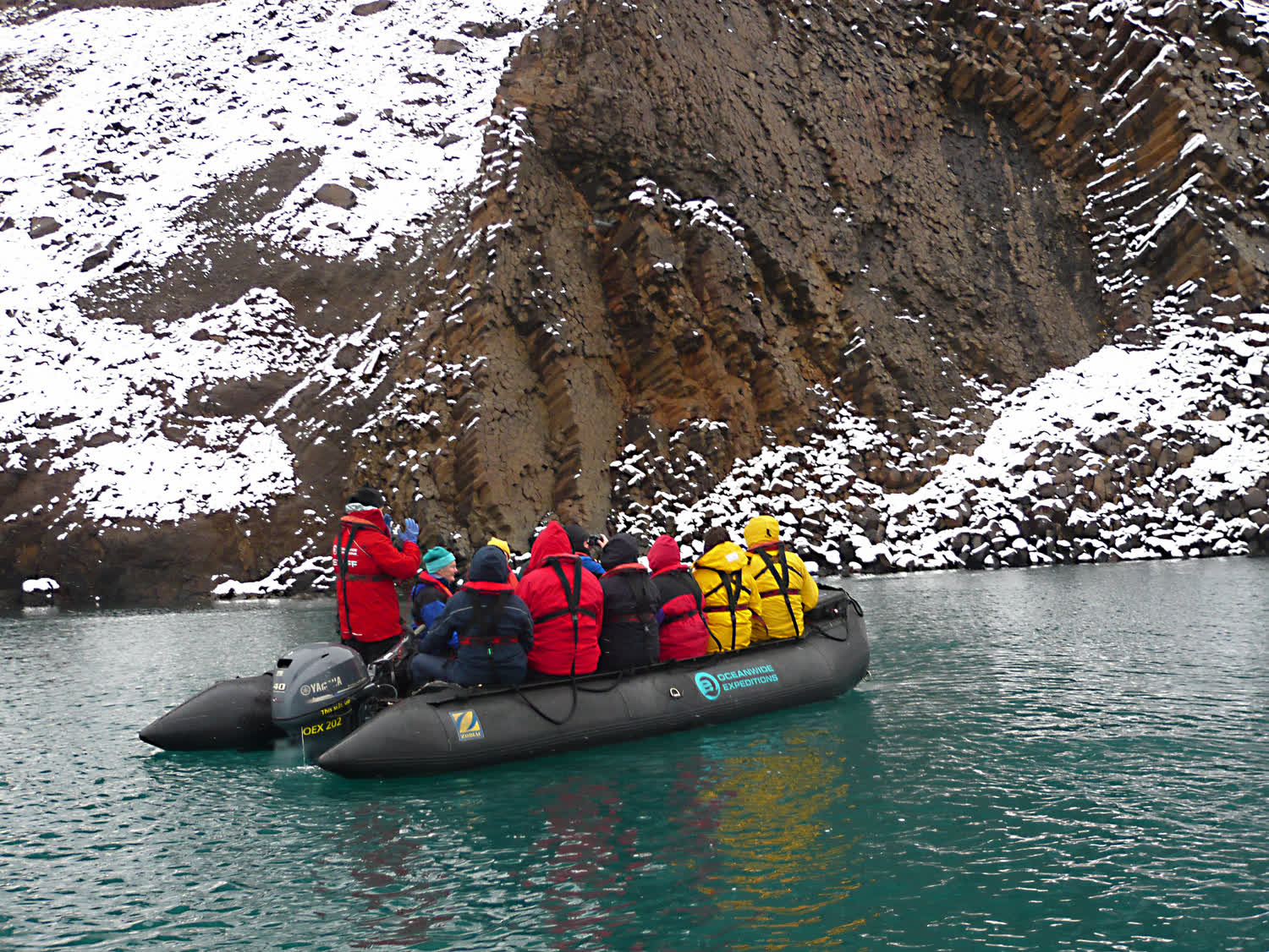 Zodiac mit Passagieren in roten Parkas vor einer Basaltwand