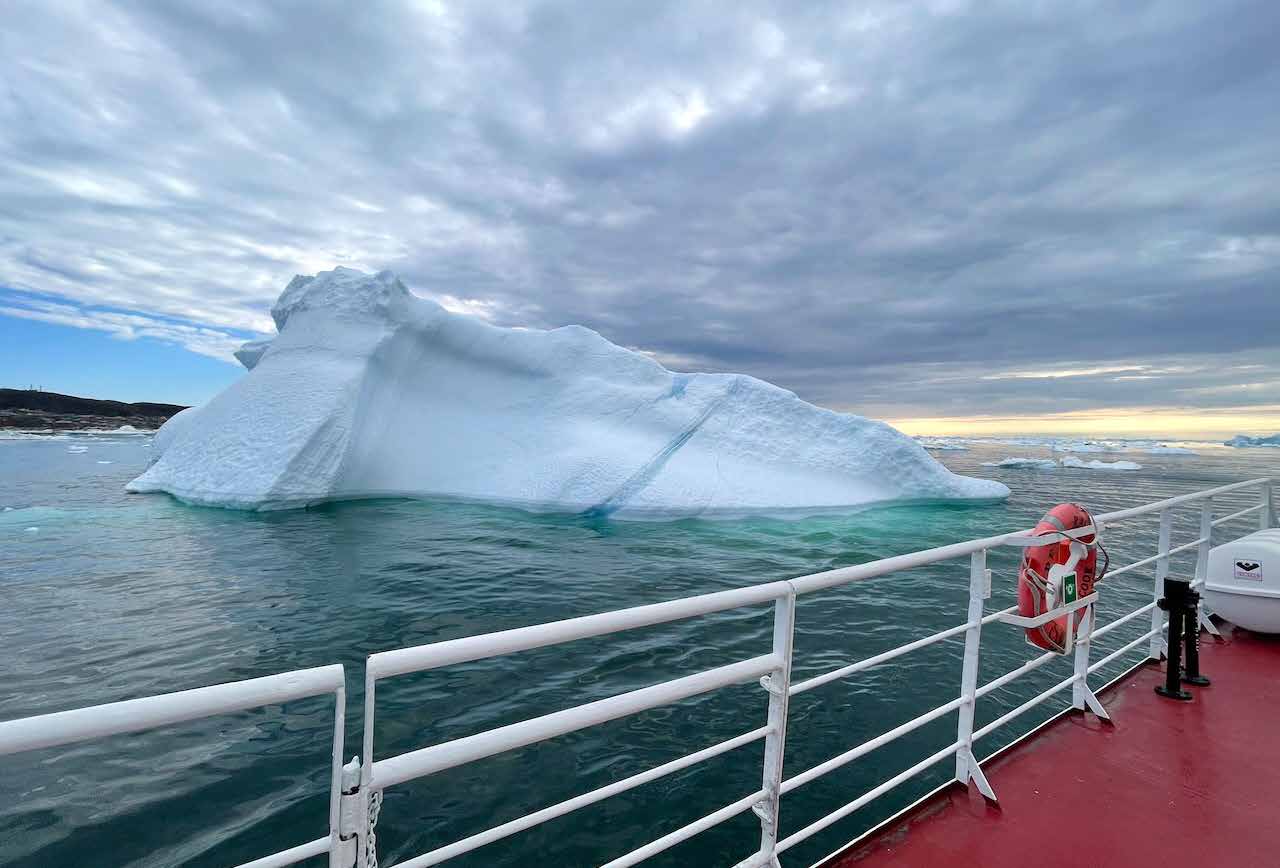 Blick über die Reling eines Schiffes auf einen großen Eisberg im Abendlicht